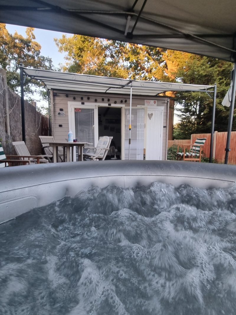 A hot tub in a back garden with a garden room behind it