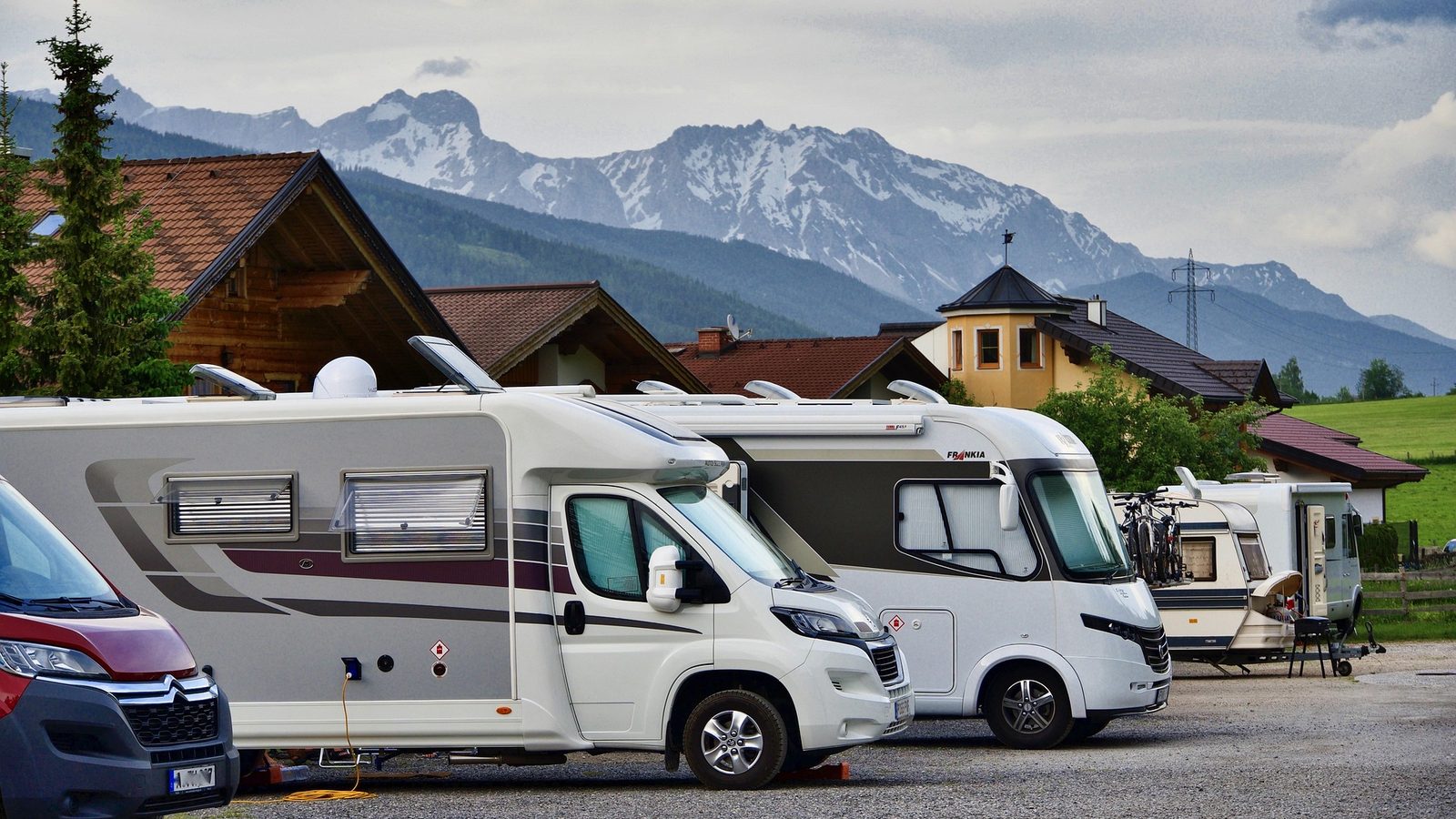 Motorhomes parked on a gravel campsite below snow-capped mountains, one carrying bikes on a rear rack